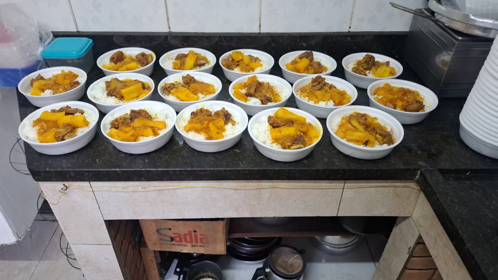 Quinze marmitas abertas e cheias de arroz, mandioca e carne em molho, alinhadas em três fileiras sobre uma bancada de granito.