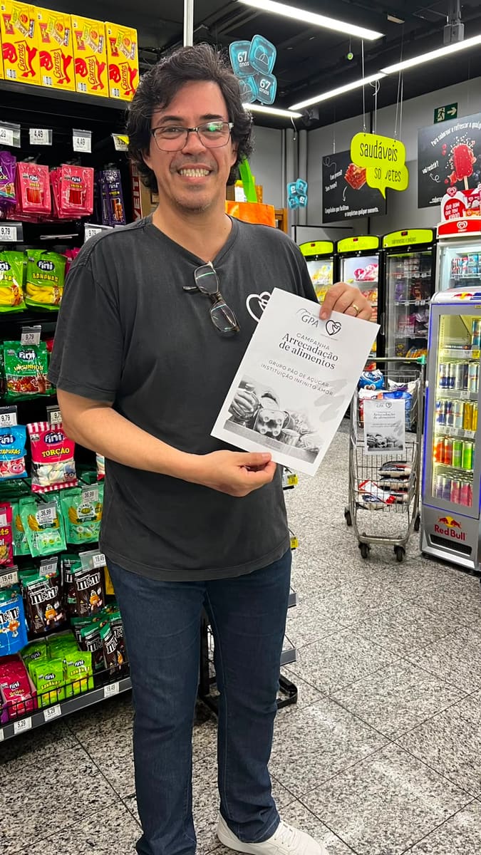 Pastor German Yaku em um supermercado, sorrindo, segurando o cartaz da Campanha de Arrecadação de Alimentos.