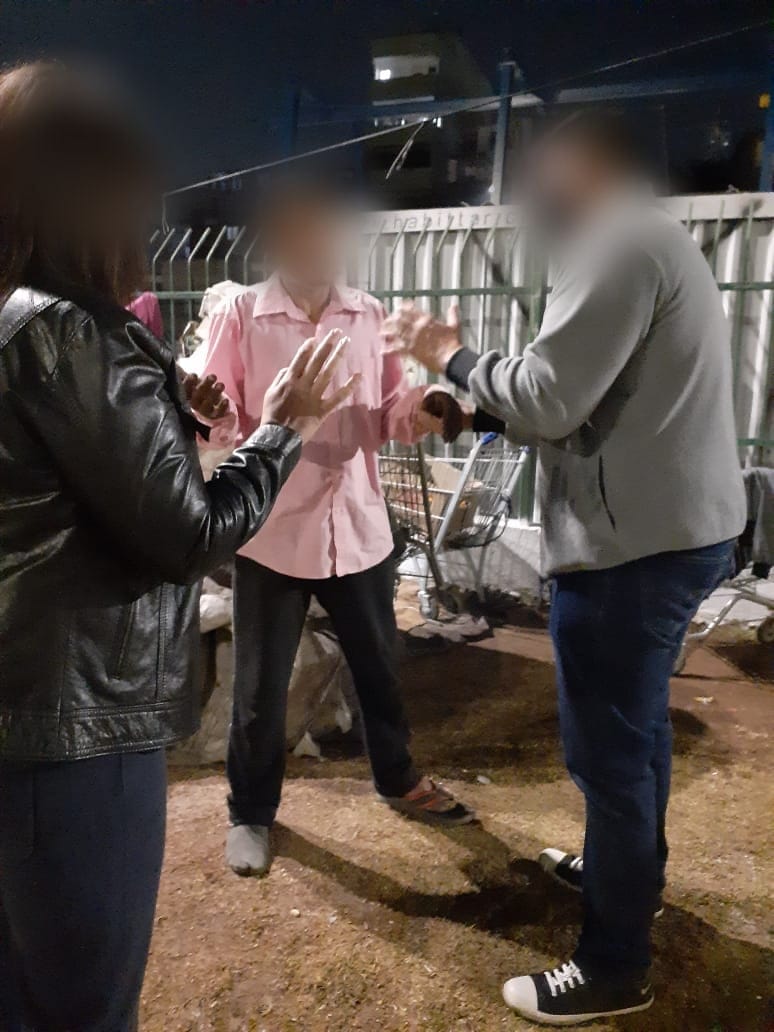 Três pessoas orando de mãos levantadas na rua, à noite, durante a entrega das marmitas. Os rostos foram desfocados.