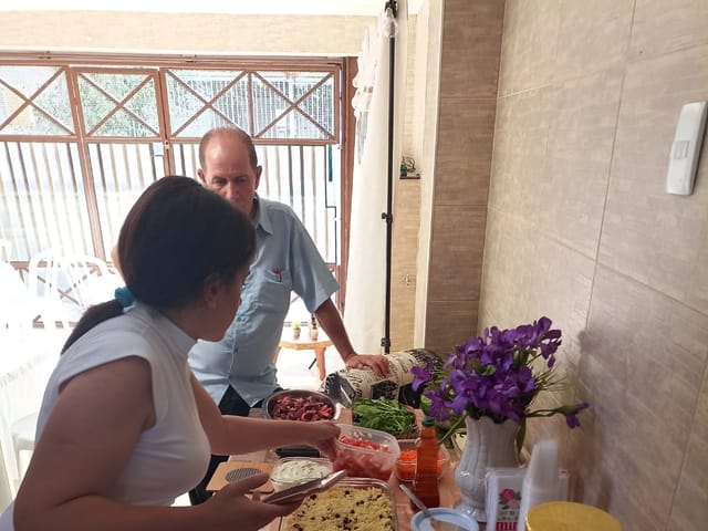Dois voluntários preparando a mesa da confraternização, com travessas de comida e um vaso de flores.
